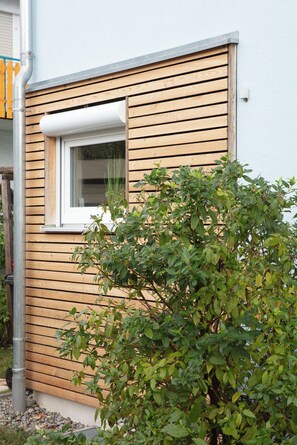 Ein Gebäude mit Holzfassade, einem Fenster mit weißem Rahmen und einem Busch vor der Tür.