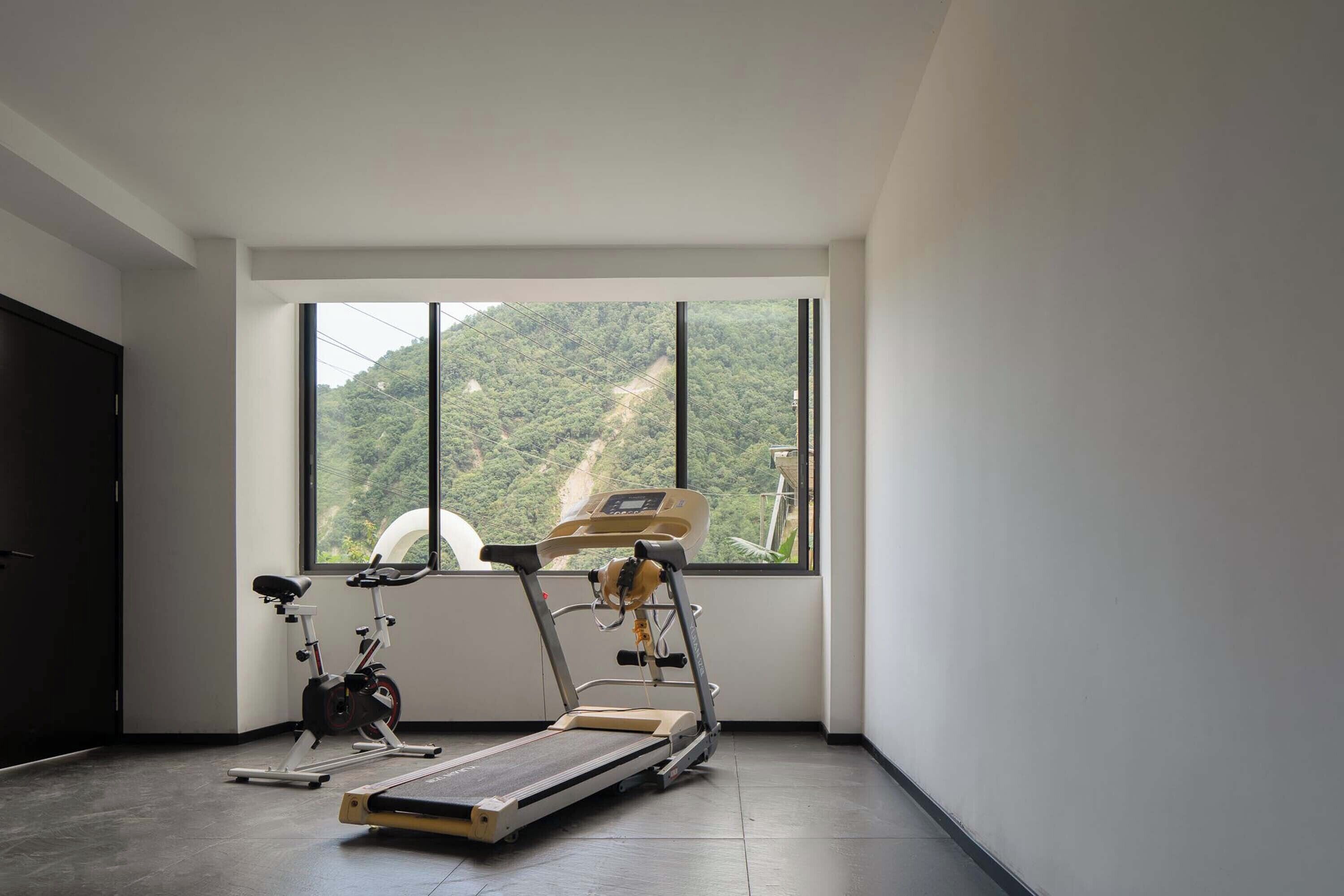 Un gimnasio con caminadora, bicicleta estática y un amplio ventanal con vistas a una ladera boscosa.