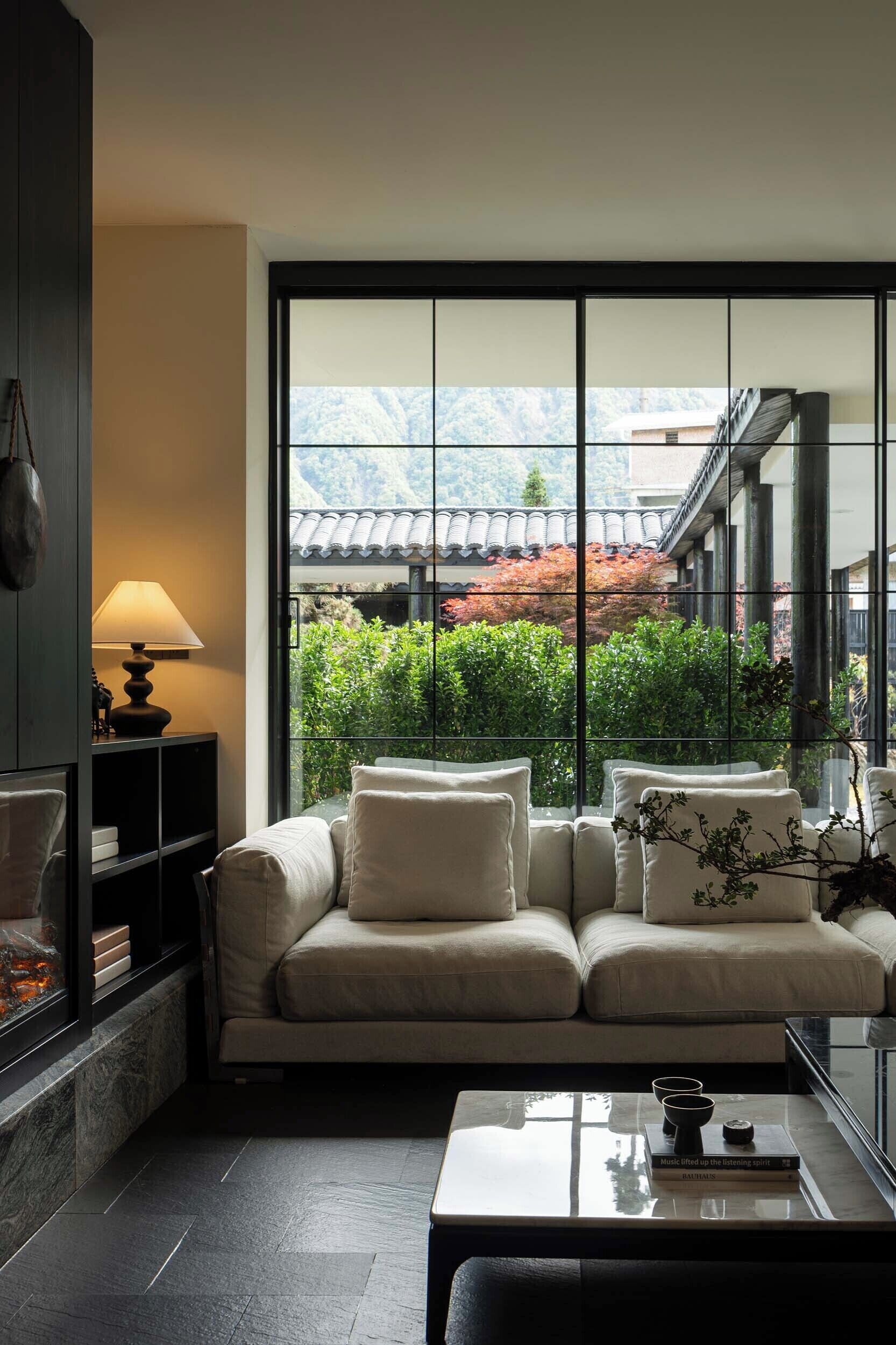 A modern living room with a sofa, a coffee table, and a lamp. A large window offers a view of a traditional building and greenery.