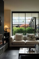 Una sala de estar moderna con un sofá, una mesita de centro y una lámpara. Un ventanal ofrece vistas a un edificio tradicional y vegetación.