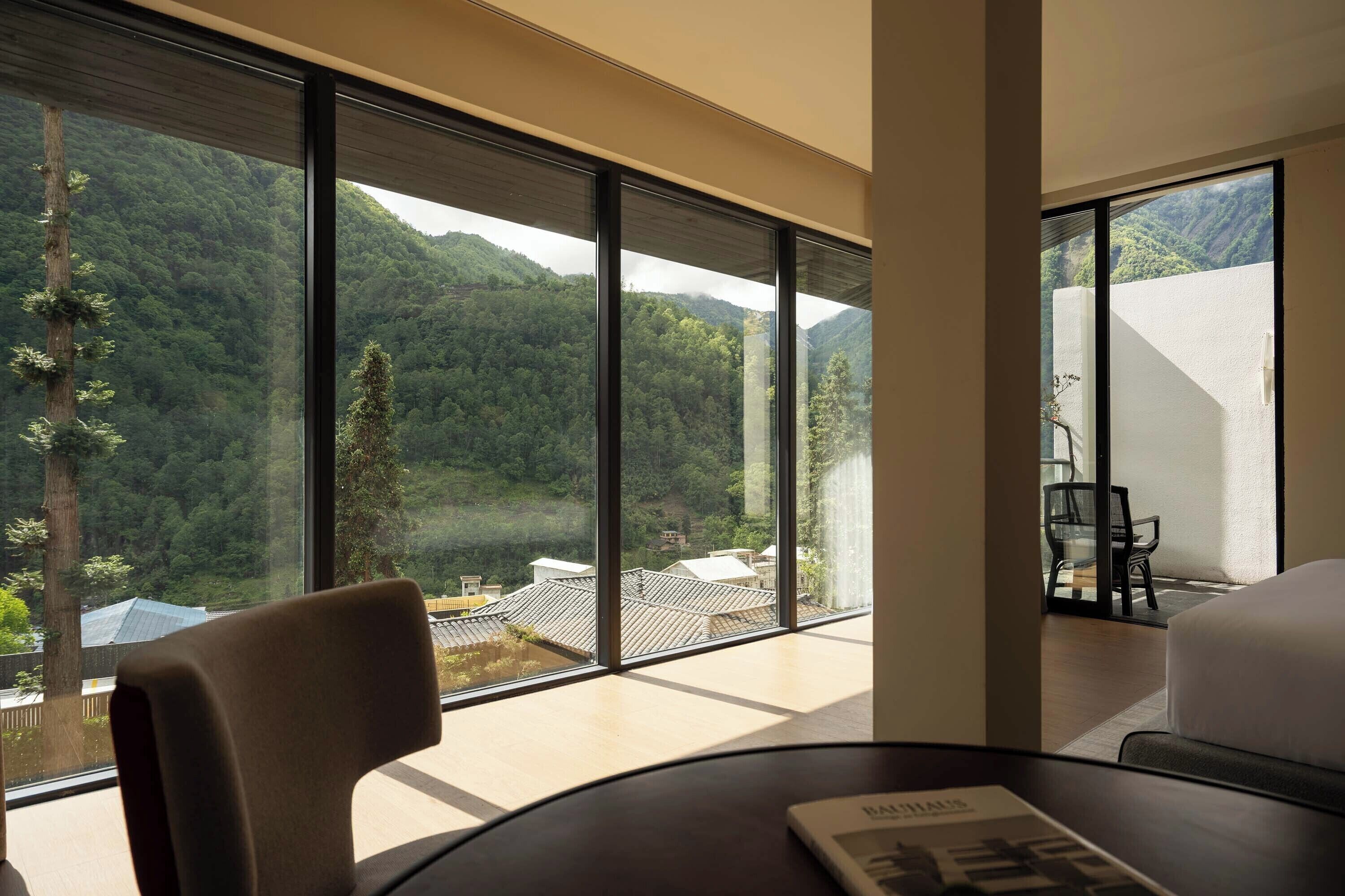 Habitación con amplios ventanales que ofrecen vistas a una montaña boscosa. Hay una mesa con una silla y una cama.