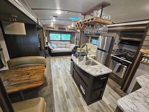Cozinha compacta de motorhome com ilha central, eletrodomésticos de aço inoxidável e área de estar com sofá e cadeiras.