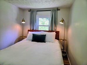 Een netjes opgemaakt bed met witte lakens en een donkroog kussen, met aan weerszijden twee nachtlampjes, in een kamer met een raam en gordijnen.