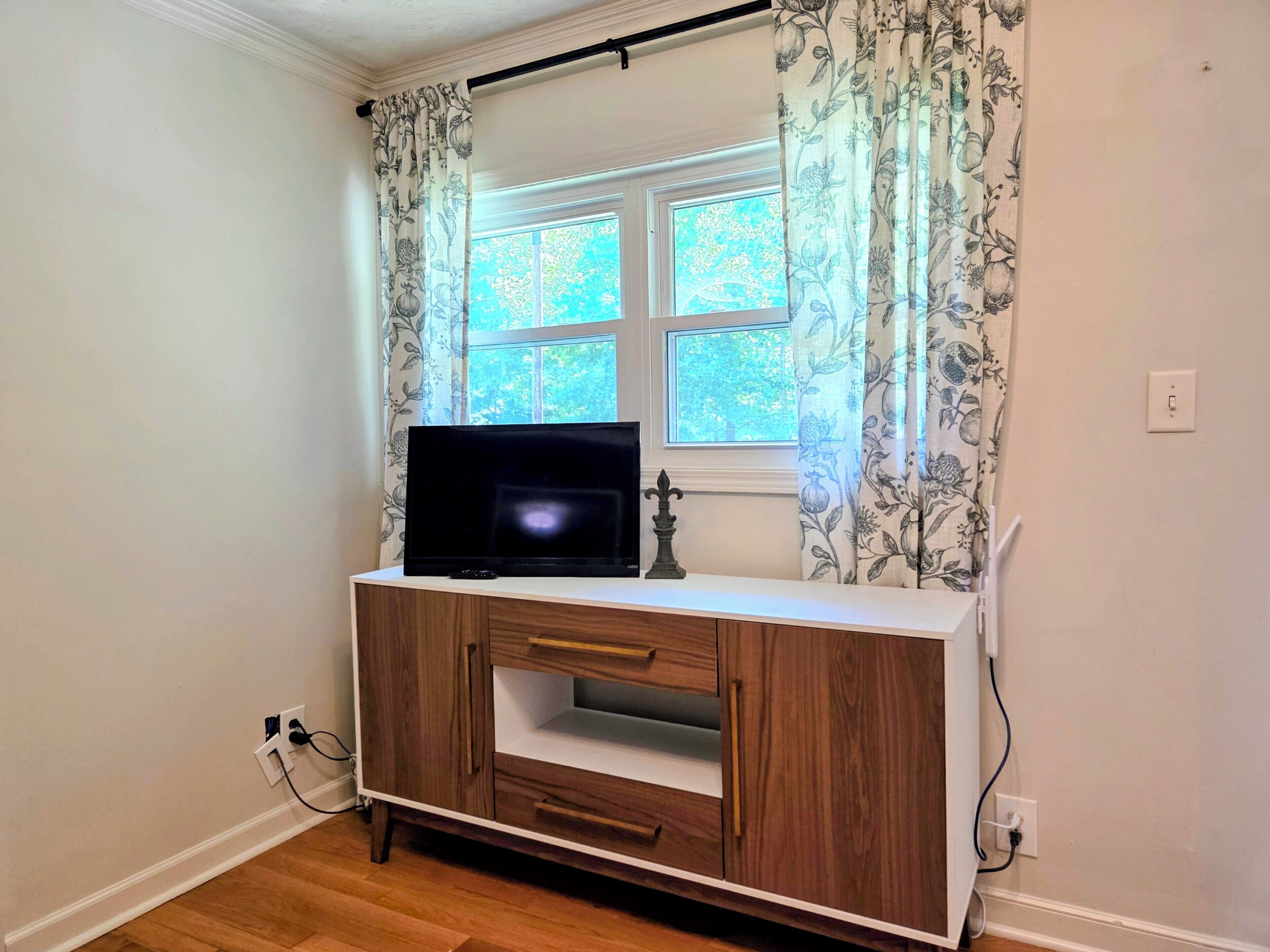A room with a television on a wooden cabinet, a window with floral curtains, and a power outlet on the wall.