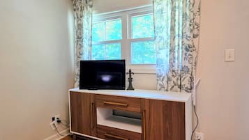 Quarto com televisão sobre um móvel de madeira, janela com cortinas florais e tomada na parede.