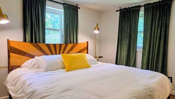 Un lit bien fait, avec une literie blanche et un seul oreiller jaune, une tête de lit en bois et des rideaux verts aux fenêtres.