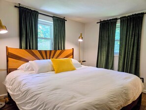 Un lit bien fait, avec une literie blanche et un seul oreiller jaune, une tête de lit en bois et des rideaux verts aux fenêtres.