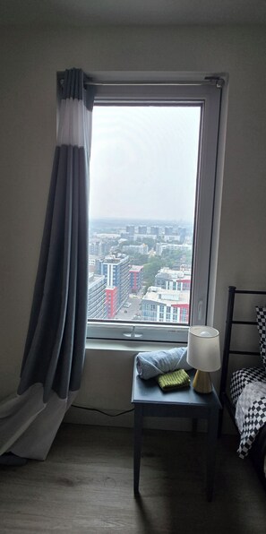 A room with a window, a small table with a lamp, a checkered bed, and a view of a cityscape.