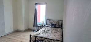 A small, single-bed room with a metal frame, a bedside table with a lamp, and a window with curtains.