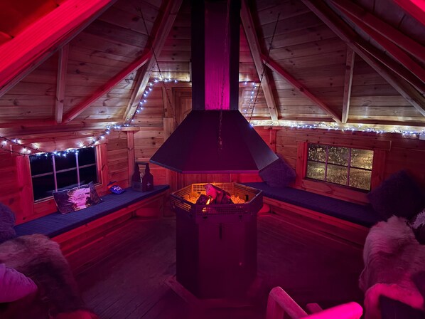 A cozy cabin interior with a central fireplace, wooden walls, and string lights.