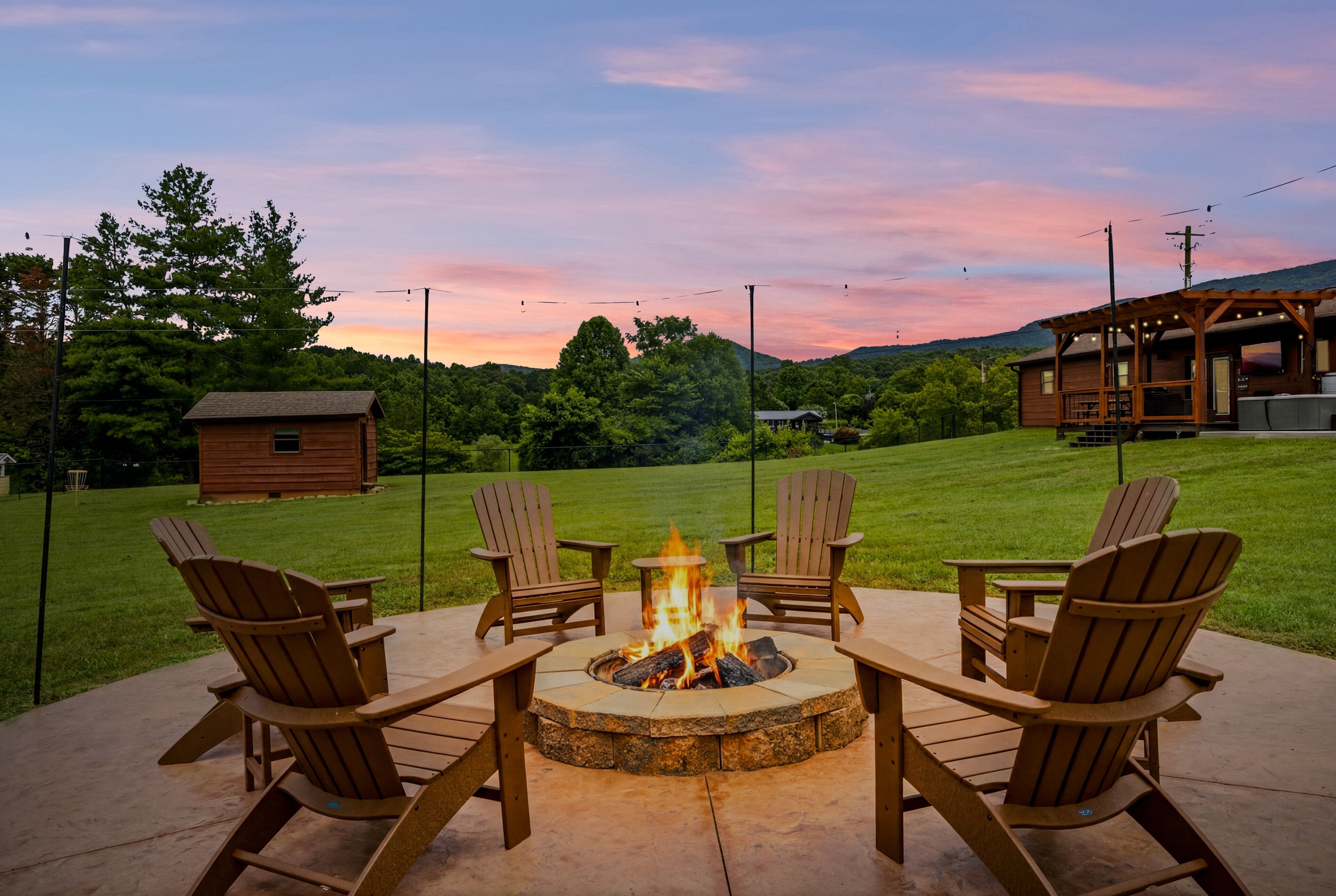 A fire pit surrounded by Adirondack chairs on a patio with a view of a wooden cabin and a grassy field.