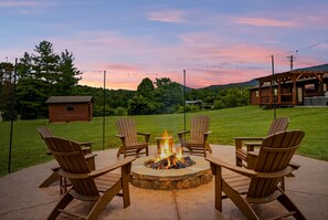 A fire pit surrounded by Adirondack chairs on a patio with a view of a wooden cabin and a grassy field. - Webster Lodge At Covemont 3 Bedroom Cabin (Sevierville)