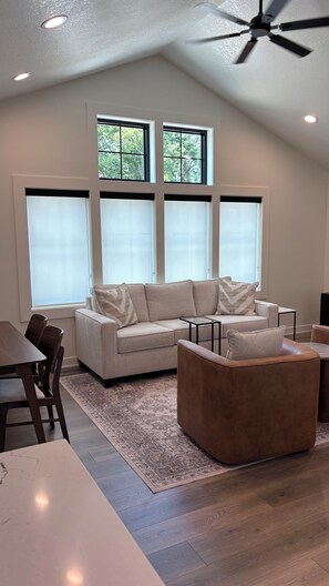 A modern living room with a sectional sofa, a brown ottoman, a glass coffee table, and a dining area with a table and chairs. - Gezellig Carriage House - The Zuid Holland Suite - Pella's Premier Retreat (PELLA)