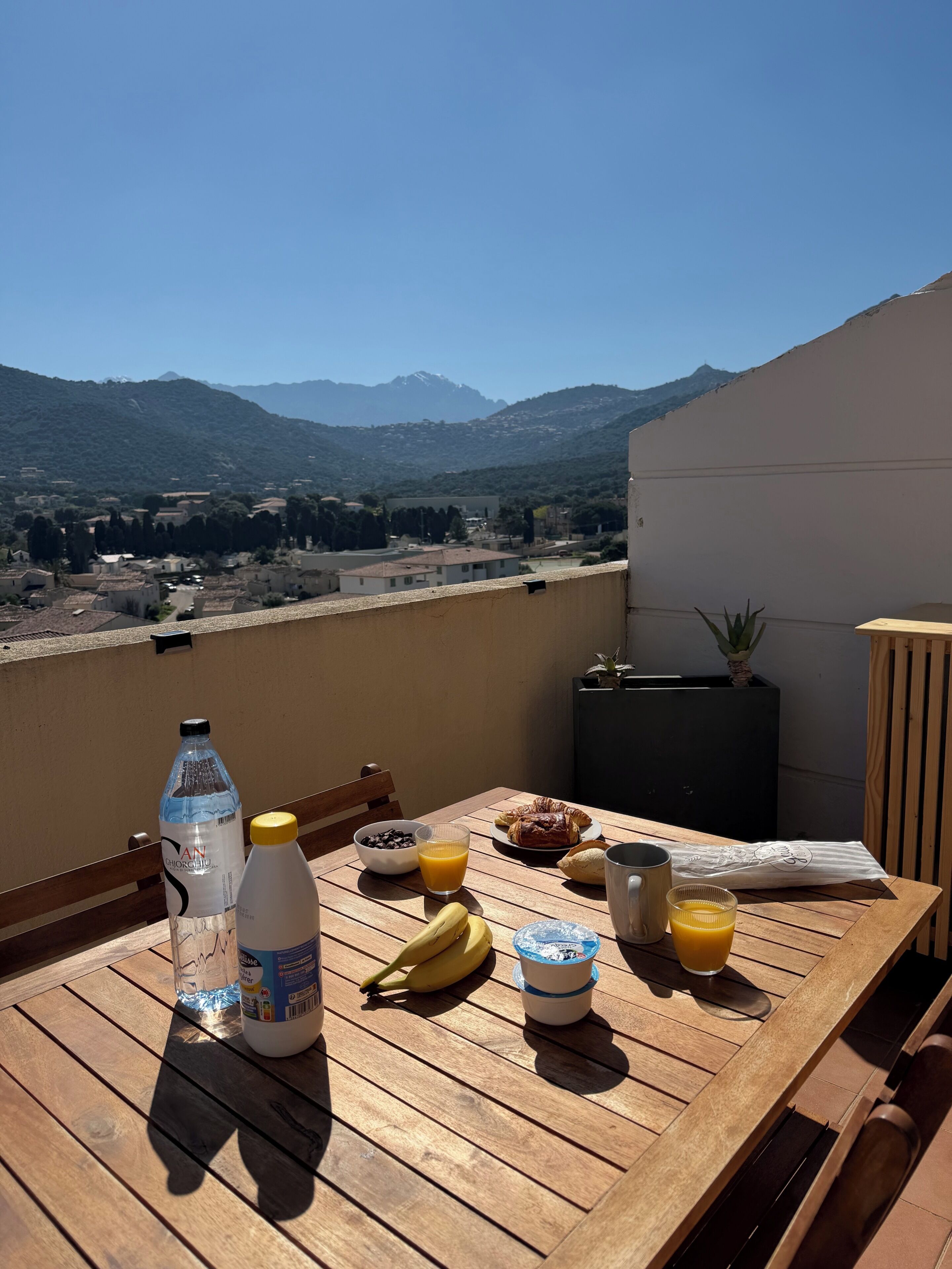 A wooden table with breakfast items, a bottle of water, a glass of orange juice, and a banana on a balcony overlooking a mountainous landscape.
