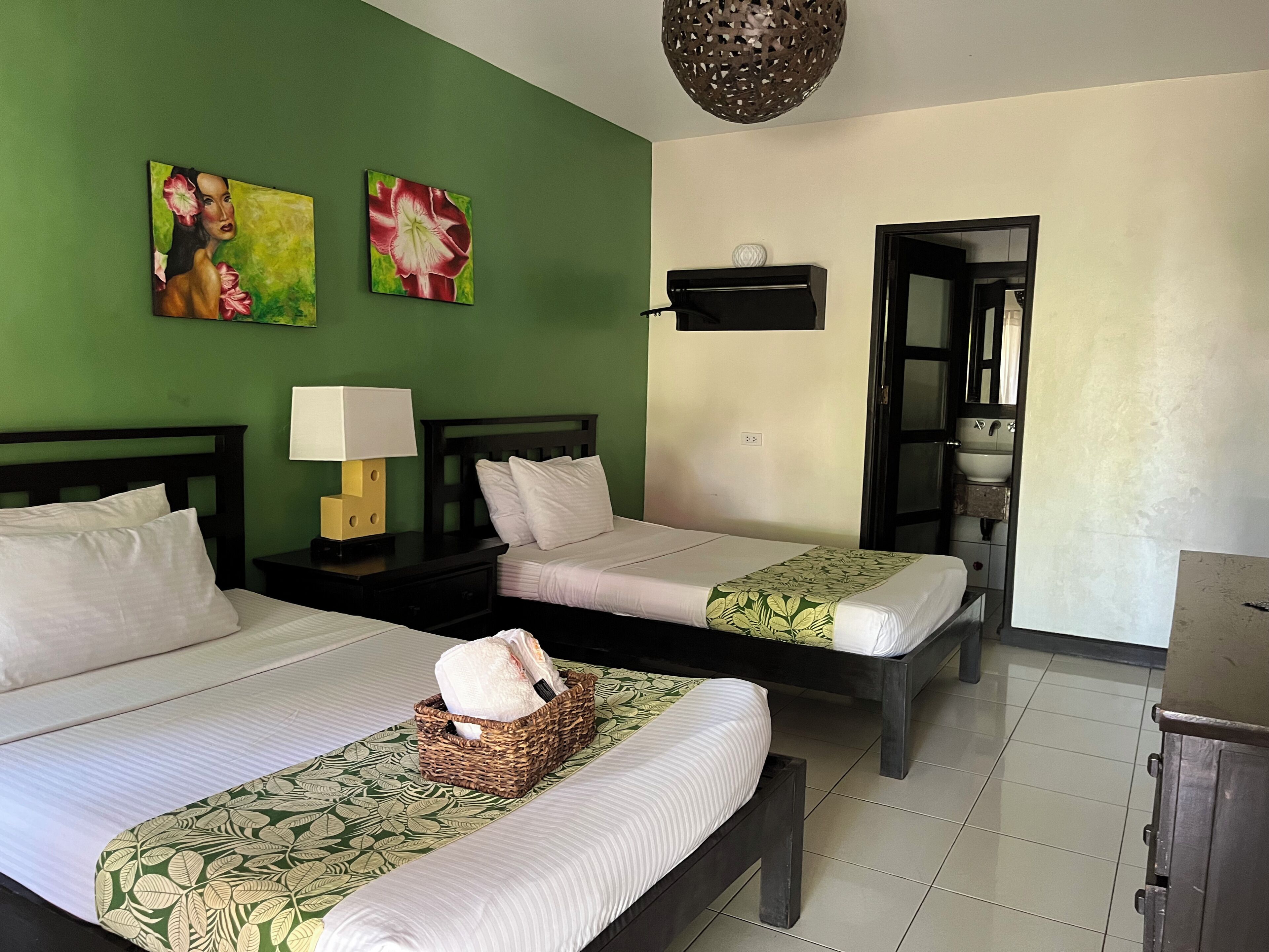 A hotel room with two beds, a nightstand with a lamp, a wall-mounted shelf, and a door leading to a bathroom.