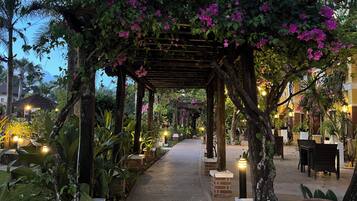 A covered pathway lined with plants and lights, leading to a building with outdoor seating.
