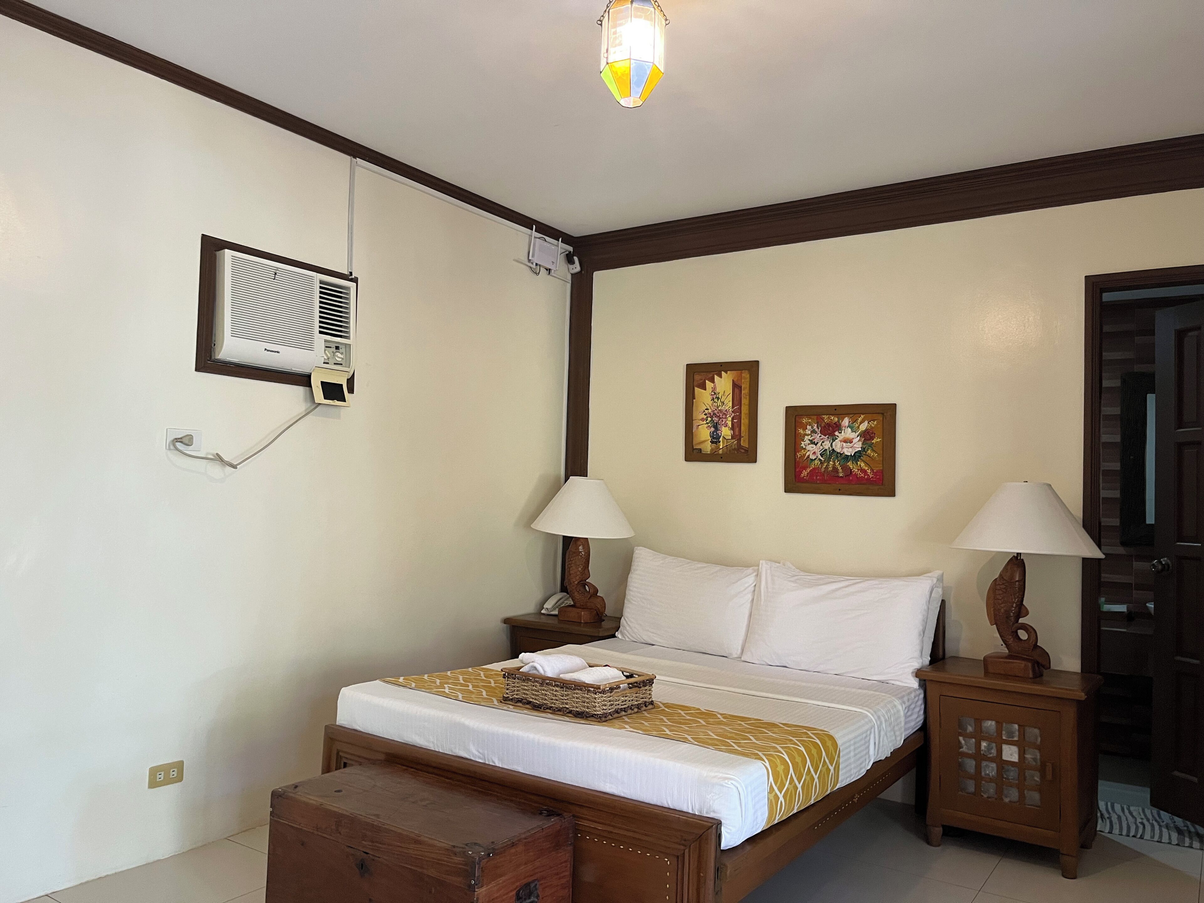 A bedroom with a bed, bedside tables, a lamp, a wall-mounted air conditioner, and a ceiling light fixture.