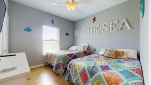 A bedroom with two beds, a ceiling fan, a window with blinds, and a wall with the text 'BY THE SEA'. - Turtlely Relaxed: 3  BR, 2  BA House in Crystal Beach, Sleeps 10 (Crystal Beach)