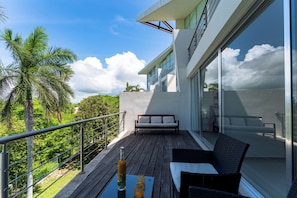 Ein Balkon mit Holzboden, einer Couch, einem Tisch mit Flasche und Glas sowie Blick auf Meer und Palmen.