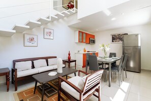 A modern living room with a sofa, coffee table, and dining area with chairs and a table. A kitchen with a refrigerator and microwave is visible in the background.