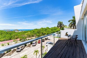 A balcony with a view of a parking area, a table with chairs, and a tropical landscape. - Main Level 2BR | Oceanview, Pool & Beach (Huatulco)