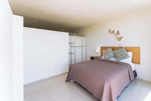A bedroom with a bed, a nightstand, a lamp, a wall-mounted shelf, and a glass door leading to a shower area. - Main Level 2-Story 2BR | Pool, Gym & Beach (Huatulco)