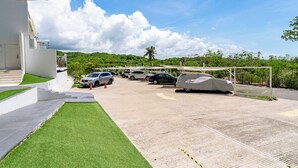 A parking area with multiple cars, a covered car, and a building with a white facade. - Main Level 2-Story 2BR | Pool, Gym & Beach (Huatulco)