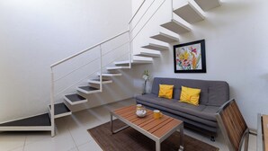 A modern living room with a staircase, a grey sofa with yellow cushions, a wooden coffee table, and a framed floral artwork on the wall. - Main Level 2-Story 2BR | Pool, Gym & Beach (Huatulco)
