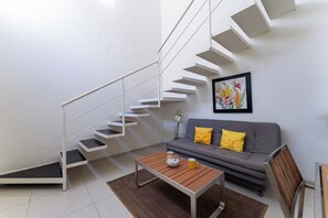 Ein modernes Wohnzimmer mit Treppe, einem grauen Sofa mit gelben Kissen, einem Holztisch und einem floralen Bild im Bilderrahmen an der Wand.