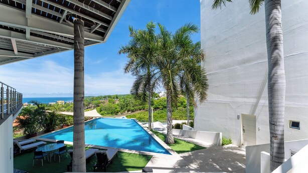A pool area with a clear view of the ocean, surrounded by palm trees and a white building. - Main Level 2-Story 2BR | Pool, Gym & Beach (Huatulco)