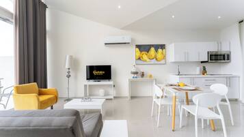 A modern living room with a sofa, a yellow armchair, a TV, a dining table with chairs, a kitchen area with cabinets and a microwave, and a painting on the wall.