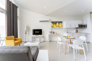 A modern living room with a sofa, a yellow armchair, a TV, a dining table with chairs, a kitchen area with cabinets and a microwave, and a painting on the wall.
