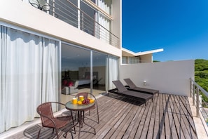 A modern outdoor balcony with a wooden deck, a glass door leading to a bedroom, and two lounge chairs.