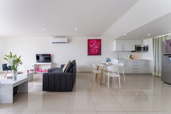 A modern living room with a sofa and a TV, a dining area with chairs and a table, and a kitchen with cabinets and appliances.
