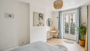 A bedroom with a bed, a desk, a chair, a hanging light fixture, a mirror, and a window with curtains. - Bel Appartement 10 Minutes Pied de la Tour Eiffel (Paris)