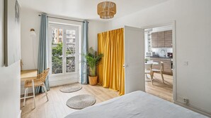 A room with a bed, a desk, a chair, a plant, and a view of a balcony and kitchen. - Bel Appartement 10 Minutes Pied de la Tour Eiffel (Paris)