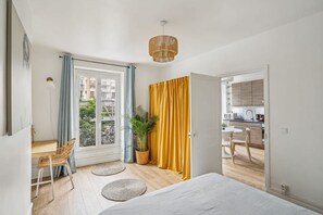 Ein Zimmer mit einem Bett, einem Schreibtisch, einem Stuhl, einer Pflanze sowie Blick auf einen Balkon und eine Küche.