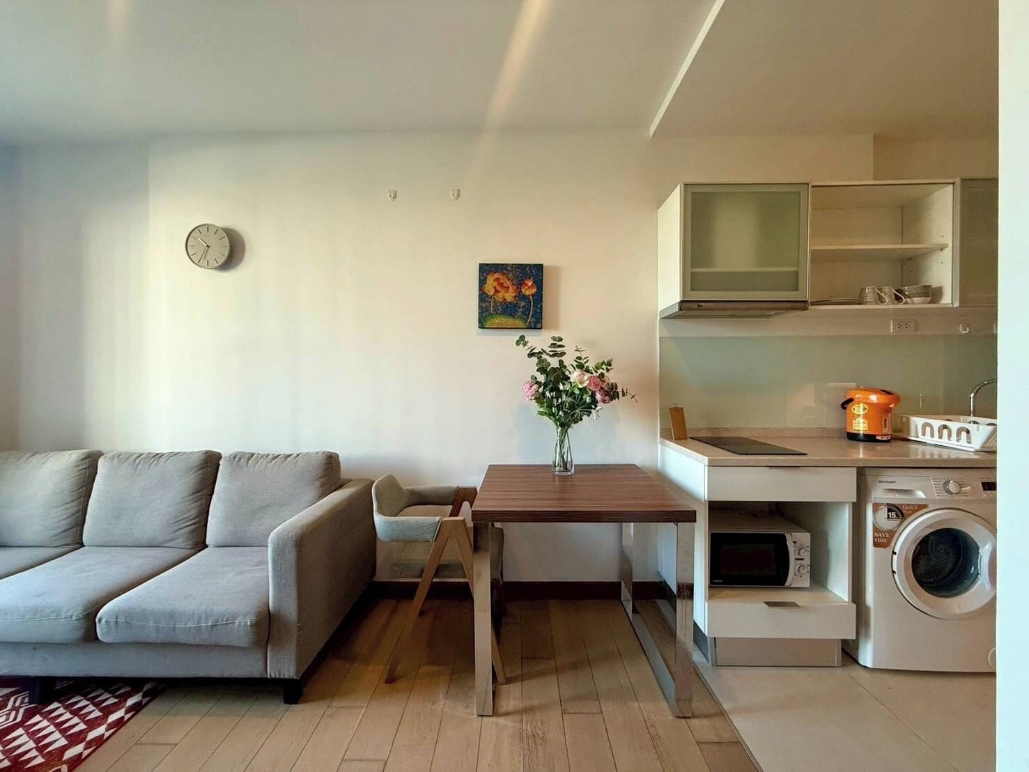 A compact living space with a sofa, a small table, a clock, a painting, a vase with flowers, a kitchen area with cabinets, a microwave, and a washing machine.