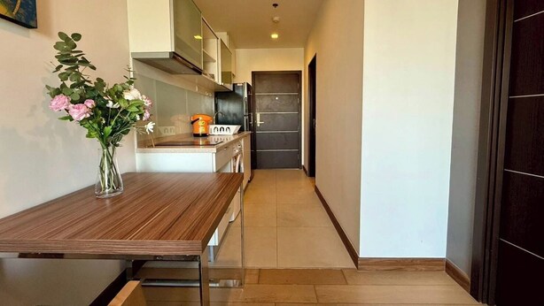 A narrow hallway with a wooden table and a vase of flowers on the left, leading to a kitchen area with a refrigerator and cabinets. - The Astra Luxe Suite-B604
Prime Location in Central Chiang Mai 
Cozy & Modern (Chang Wat Chiang Mai)