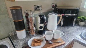 A kitchen counter with a coffee maker, a kettle, a plate with cookies, and a mug rack. - Cabin Sleeps 4 With Indoor Pool (Tywyn)