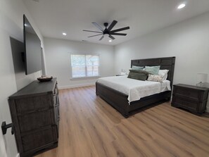 Une chambre à coucher avec un lit, un ventilateur de plafond, une télévision, une fenêtre avec des stores et un plancher en bois.