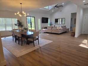Un salon spacieux avec un plafond haut, un coin repas avec une table et des chaises en bois, et une grande fenêtre qui laisse entrer la lumière naturelle.