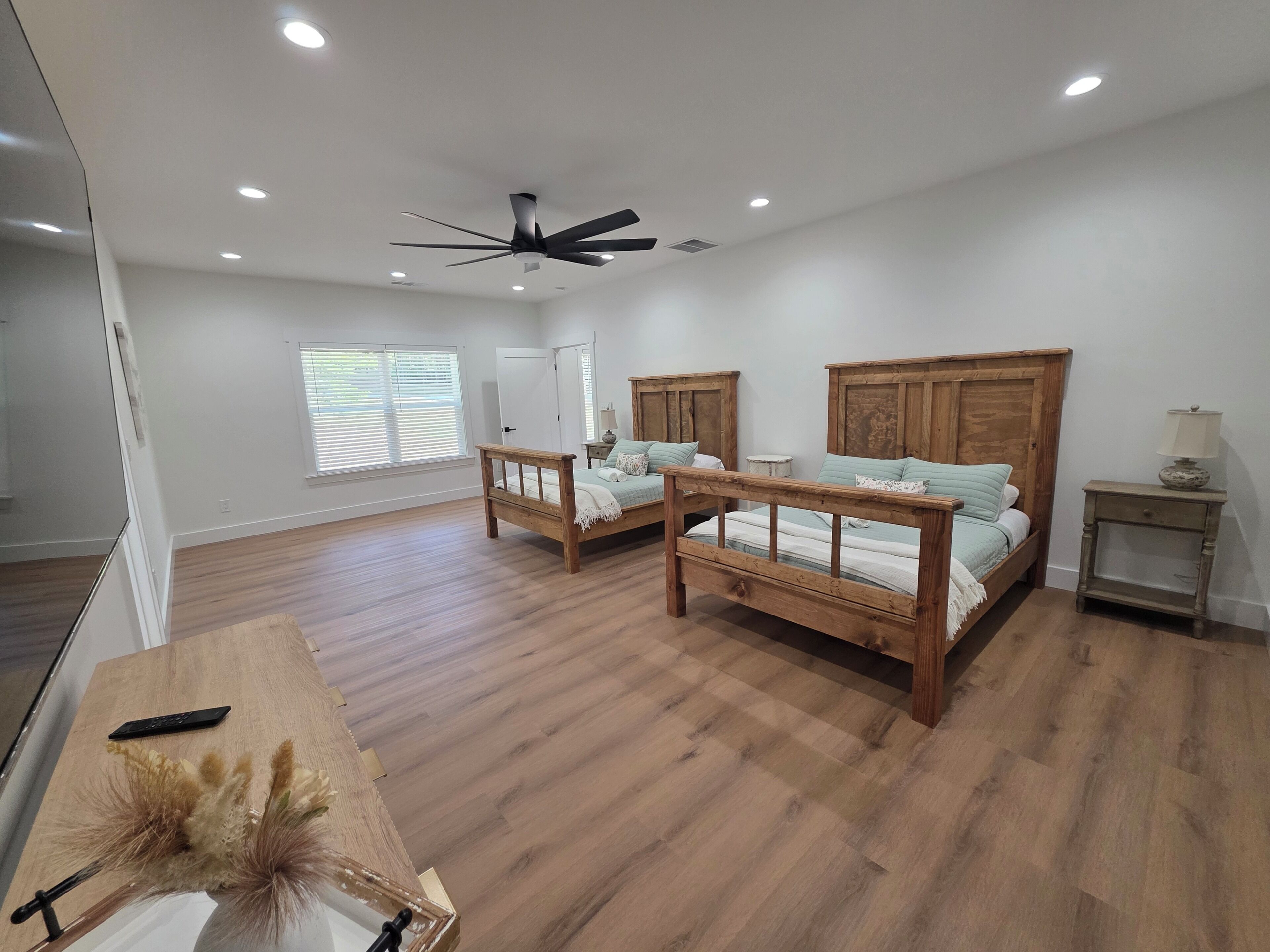A spacious bedroom with two wooden beds, a ceiling fan, and a large window with blinds.