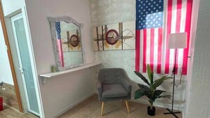 Una habitación con suelo de madera, un sillón gris, una planta en maceta, una pared con una bandera estadounidense y un espejo con marco decorativo.