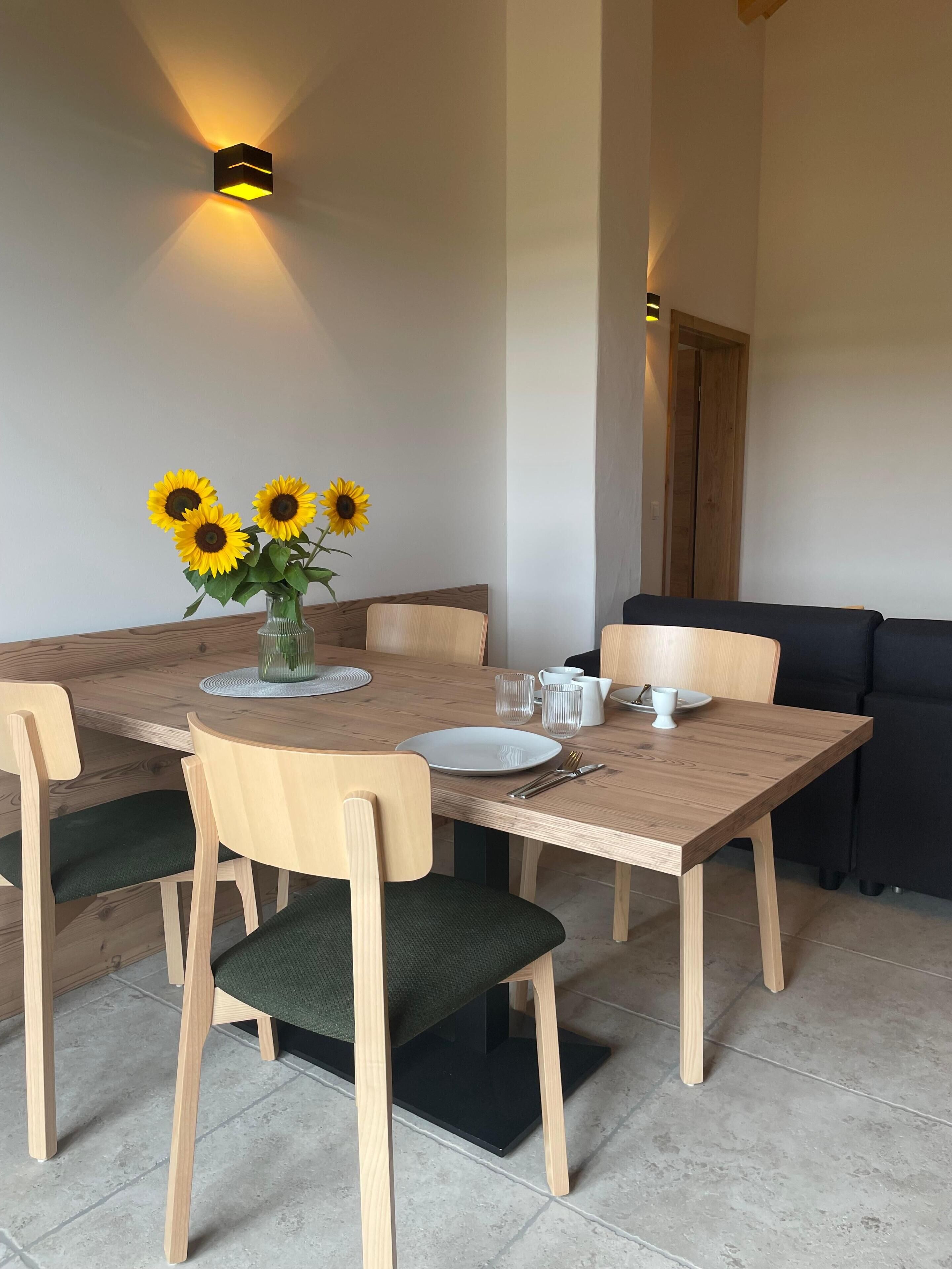 A dining area with a wooden table, chairs, a vase with sunflowers, and a wall-mounted light fixture.