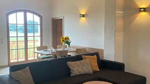 A modern living room with a dark-colored sectional sofa, a round wooden coffee table, and a large window with a view of a patio. - Bavasol – Your Temporary Bavarian Retreat (Schechen)