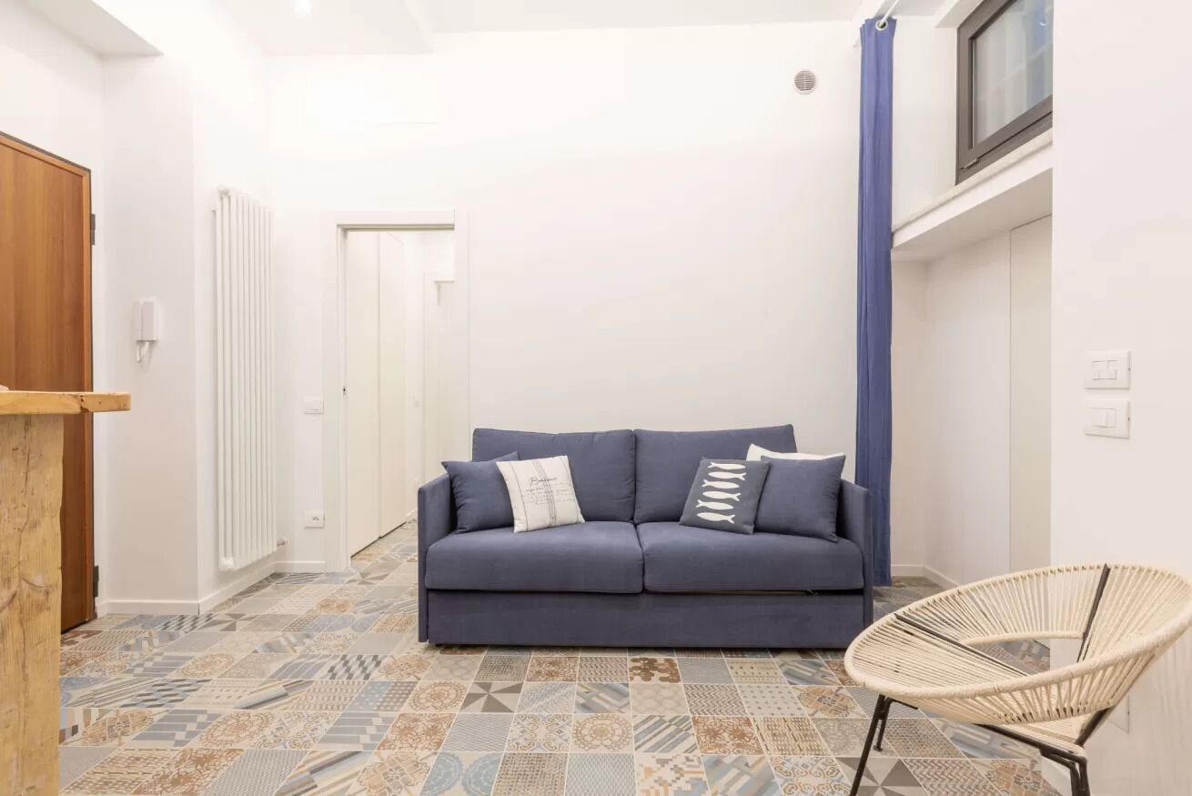 Una habitación con un sofá azul, un sillón de mimbre, baldosas de suelo estampadas, un radiador blanco y una puerta.