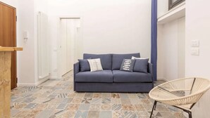 Una habitació amb un sofà blau, una cadira de vimet, terres de rajoles estampades, un radiador blanc i una porta.