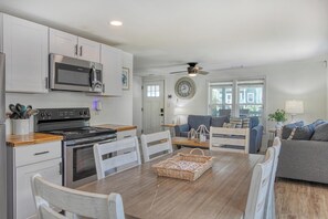 A modern kitchen with stainless steel appliances, a dining area with white chairs, and a living room with a gray sofa. - Cheeky Crab A - 2 Bedroom Coastal Cottage Steps from the Beach (Garden City Beach)