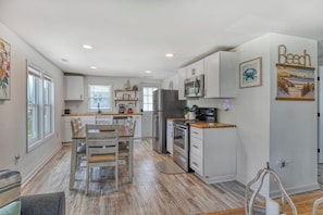 A modern kitchen with white cabinetry, a dining area with wooden chairs, and a kitchen island. - Cheeky Crab A - 2 Bedroom Coastal Cottage Steps from the Beach (Garden City Beach)